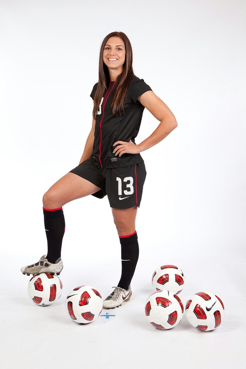 Alex Morgan wall art print features a smiling soccer player in a black number 13 kit posed among white and red balls.
