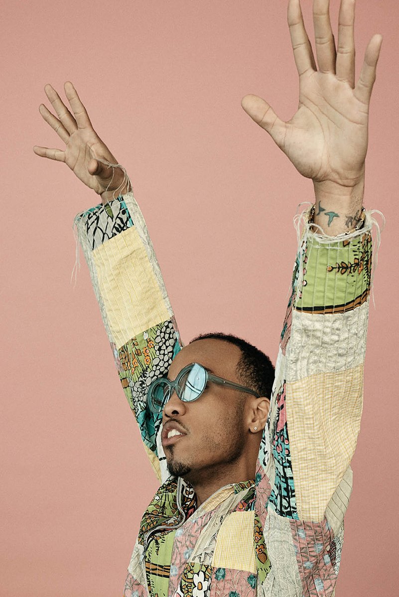 Anderson .Paak poster portrait with arms raised, blue-tinted sunglasses and a patchwork jacket against a dusty pink background.