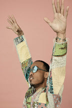 Anderson .Paak poster portrait with arms raised, blue-tinted sunglasses and a patchwork jacket against a dusty pink background.