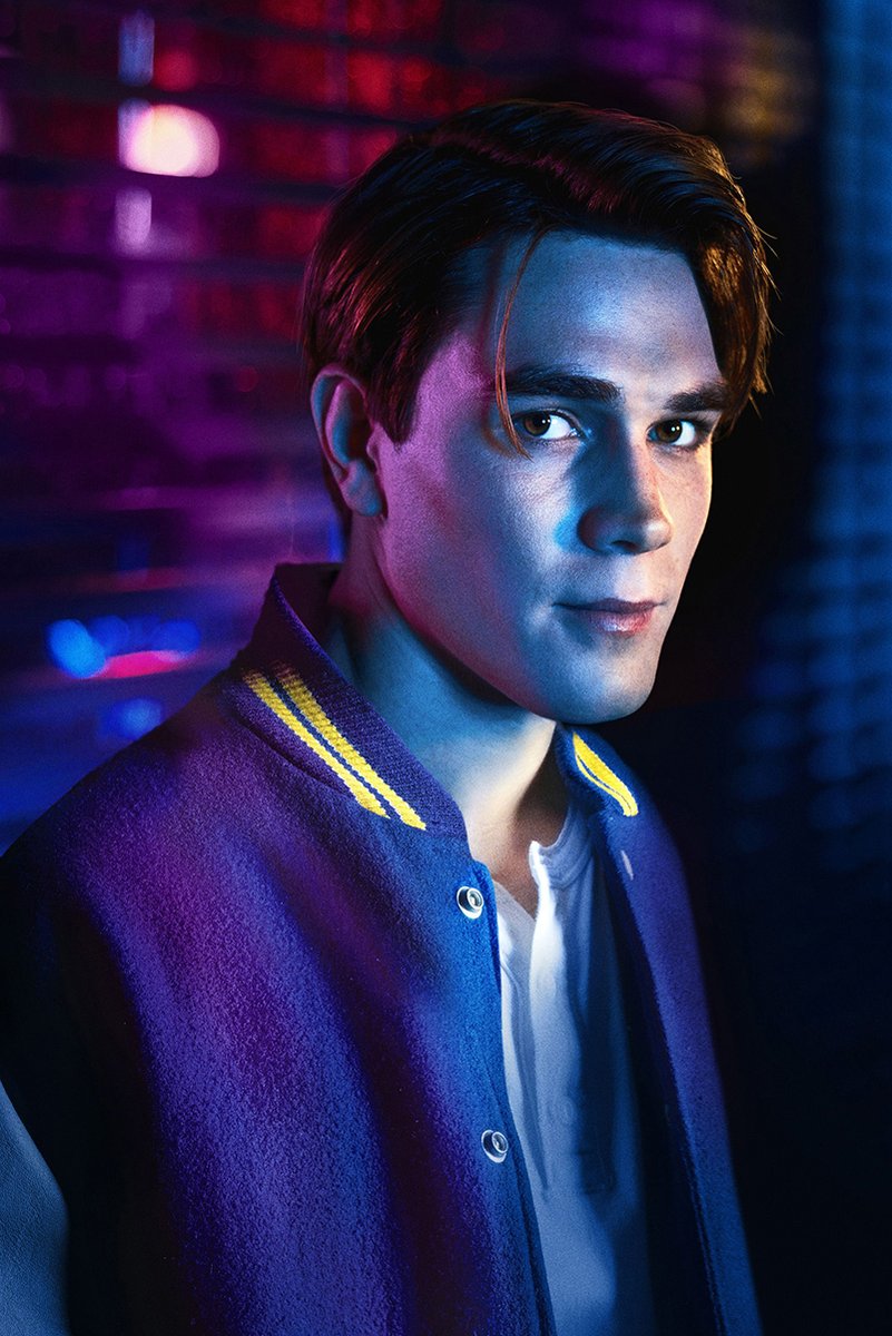 Archie Andrews poster presents a moody chest-up portrait in a blue jacket with yellow trim and a blurred neon brick backdrop.