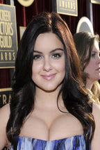 Ariel Winter poster portrait at an awards event, smiling in a strapless blue patterned dress before red curtains and signs.