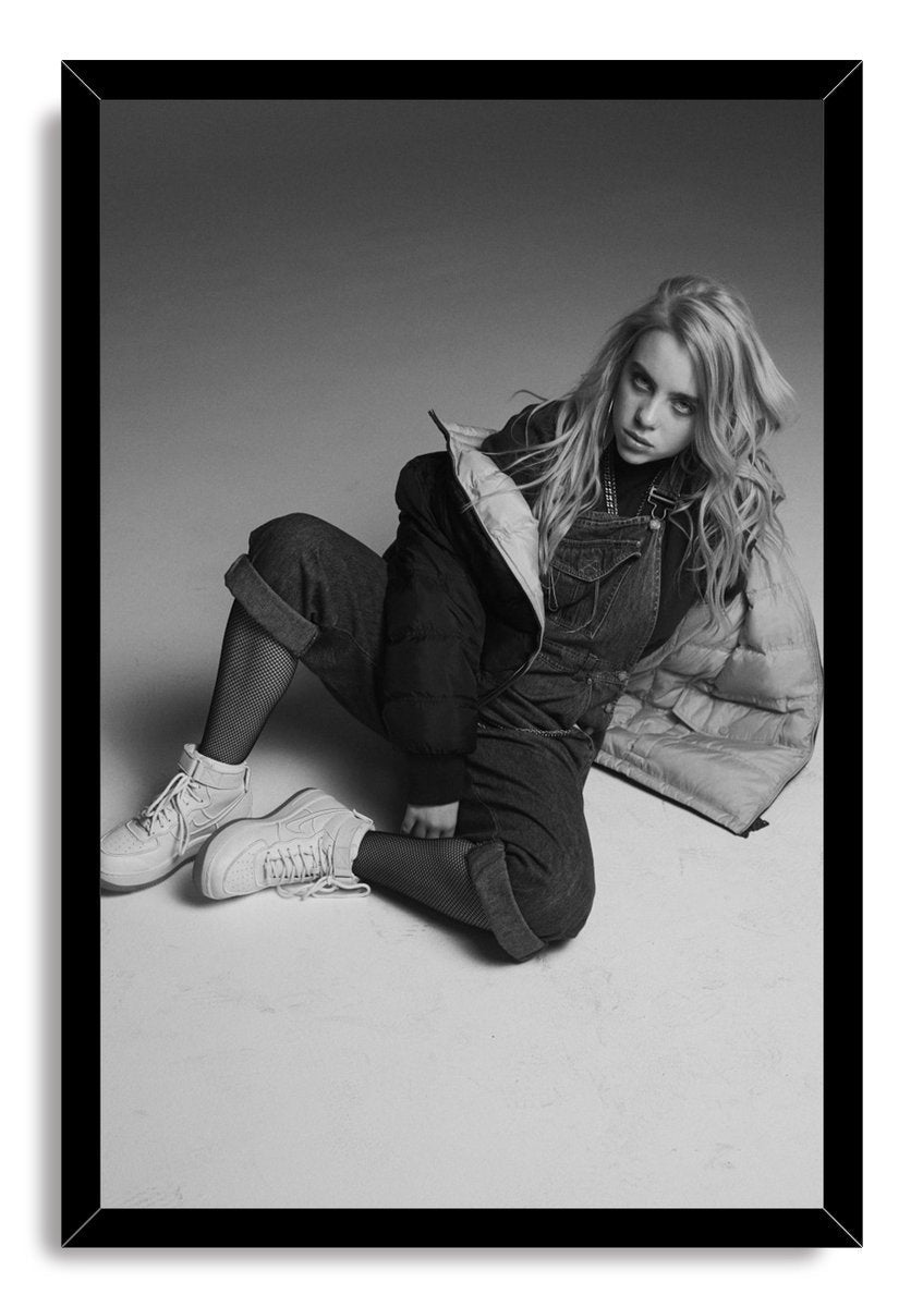 Billie Eilish framed wall art print features a monochrome floor portrait of her in denim overalls, a puffer jacket, fishnet tights, and sneakers.