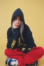 Billie Eilish wall art print portrays the singer seated cross-legged in a black hoodie and red pants against yellow.