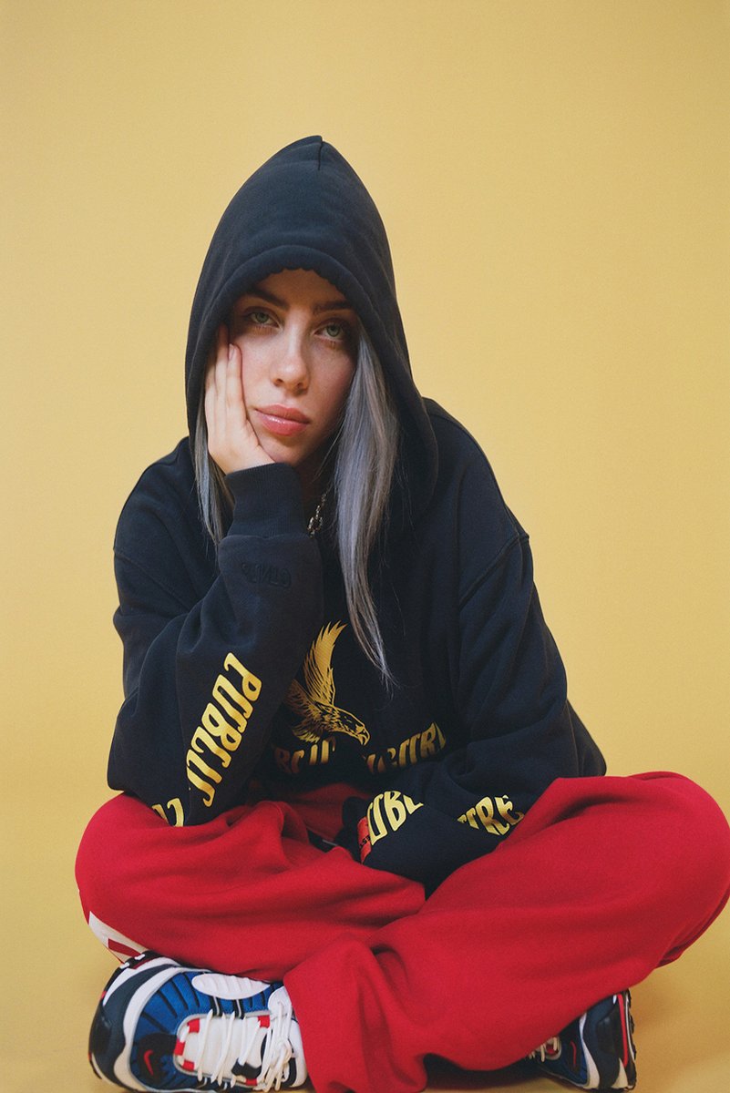 Billie Eilish wall art print portrays the singer seated cross-legged in a black hoodie and red pants against yellow.