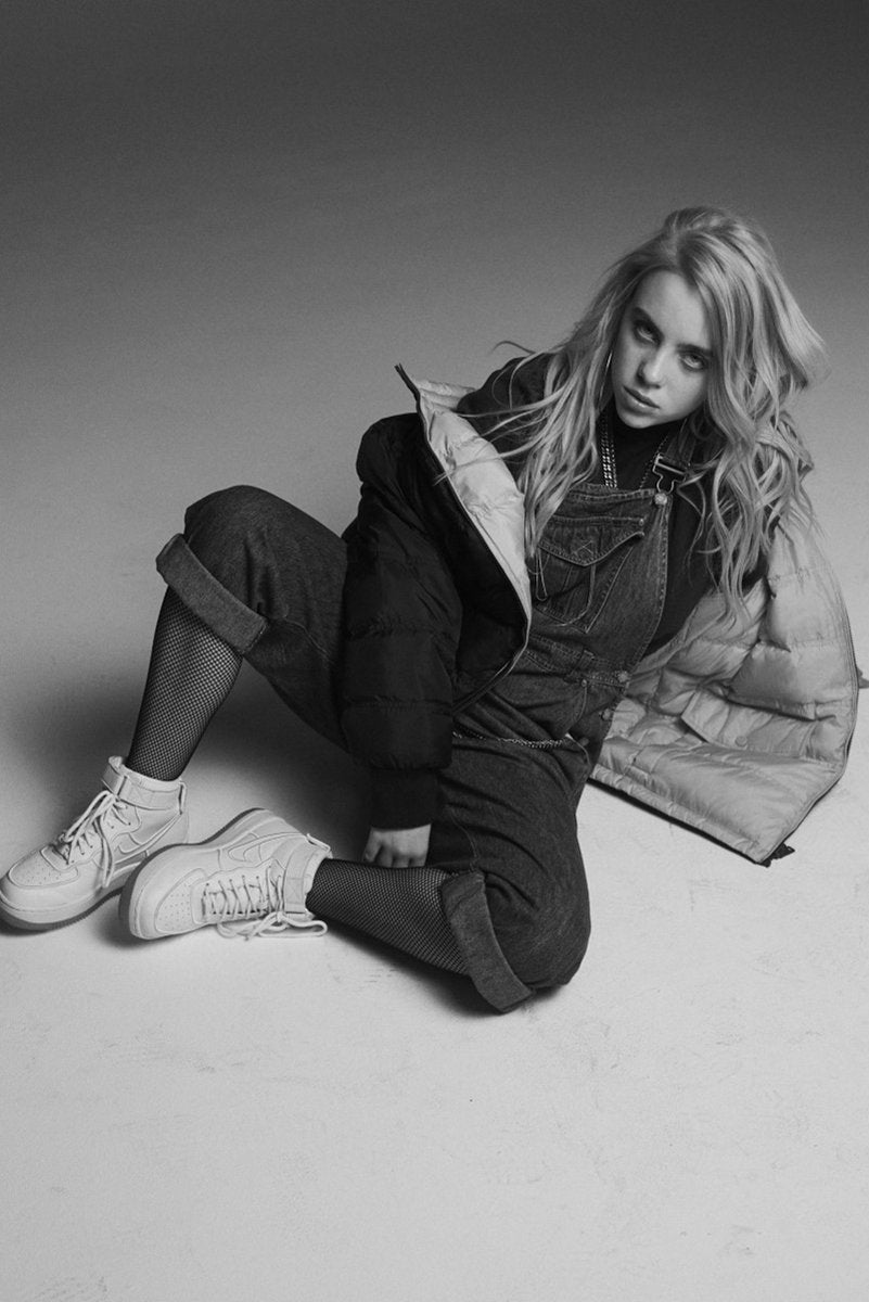 Billie Eilish wall art print in black and white, seated on the floor in overalls, a puffer jacket, fishnet tights, and sneakers.