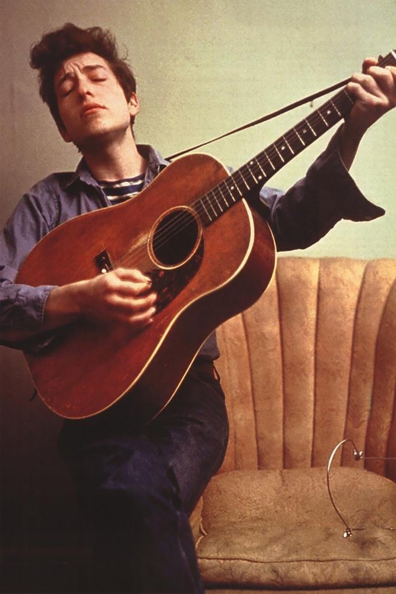 Bob Dylan poster features the musician with closed eyes strumming a brown acoustic guitar beside a tan upholstered chair.