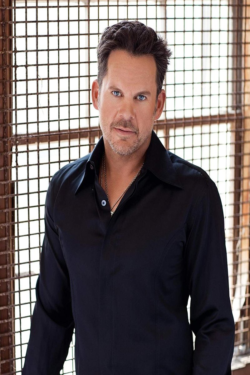 Male celebrity poster portrait with blue eyes in a dark open-collar shirt, posed before a sunlit metal grid window.