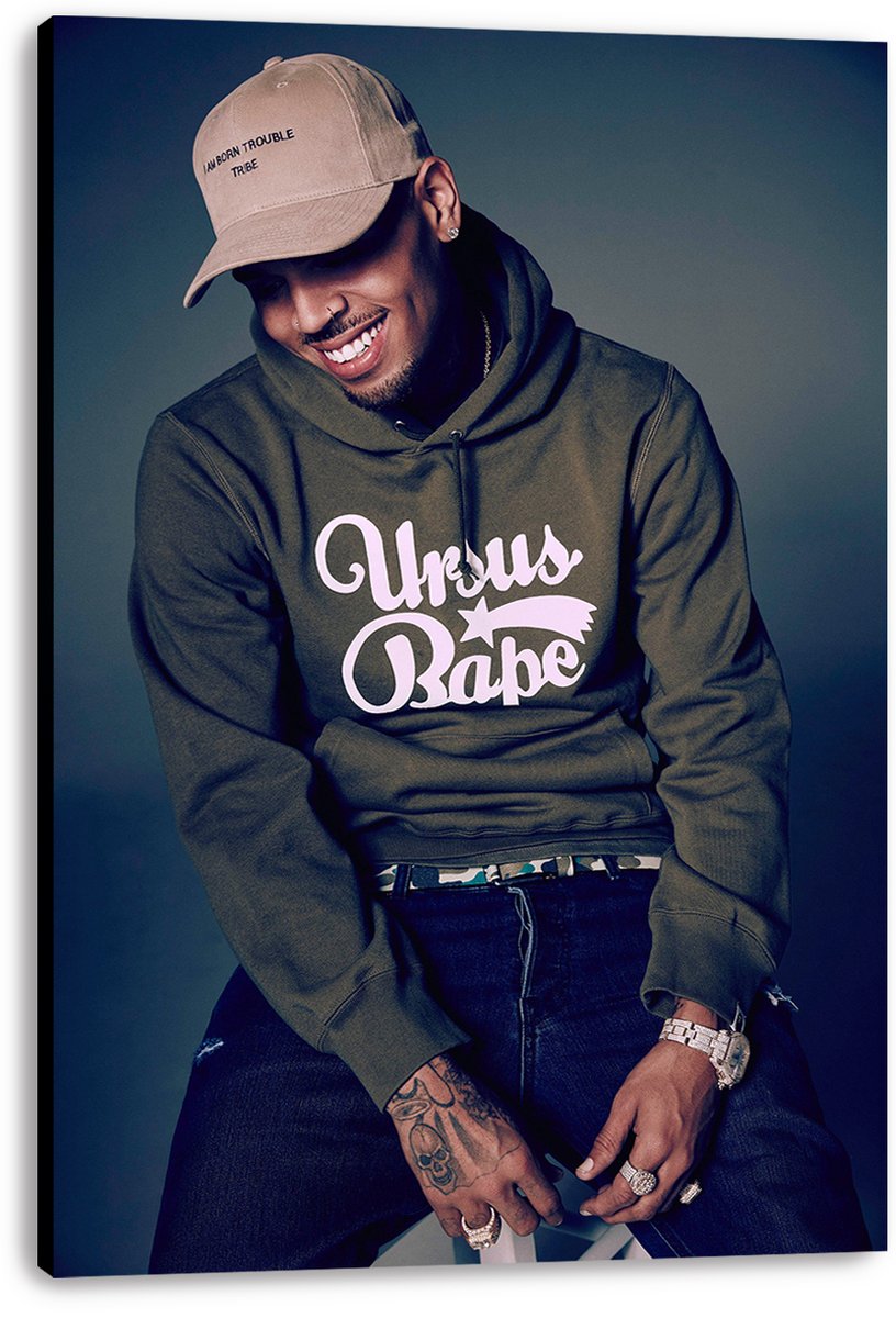 Chris Brown canvas wall art print featuring the seated singer smiling in a tan cap, green hoodie, jeans, rings, and watch.