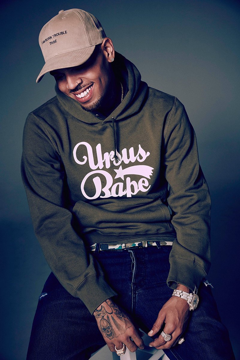 Chris Brown poster portrait of the smiling singer in a tan cap and green graphic hoodie against a dark blue-gray backdrop.