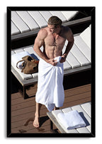Chris Hemsworth framed wall art print portrays the shirtless actor adjusting a white towel on a wooden deck beside white lounge chairs.
