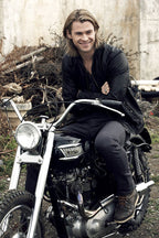 Chris Hemsworth wall art print portrays the smiling actor in a black jacket seated on a black vintage motorcycle outdoors.