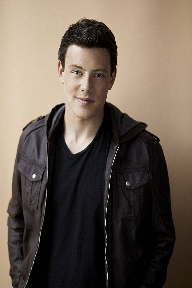 Cory Monteith poster portrait with a soft smile, dark hair, black shirt, and brown jacket against a warm beige background.