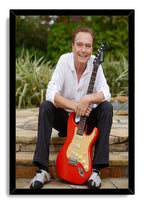 David Cassidy framed wall art print portrays the smiling musician seated with a red electric guitar against lush greenery.