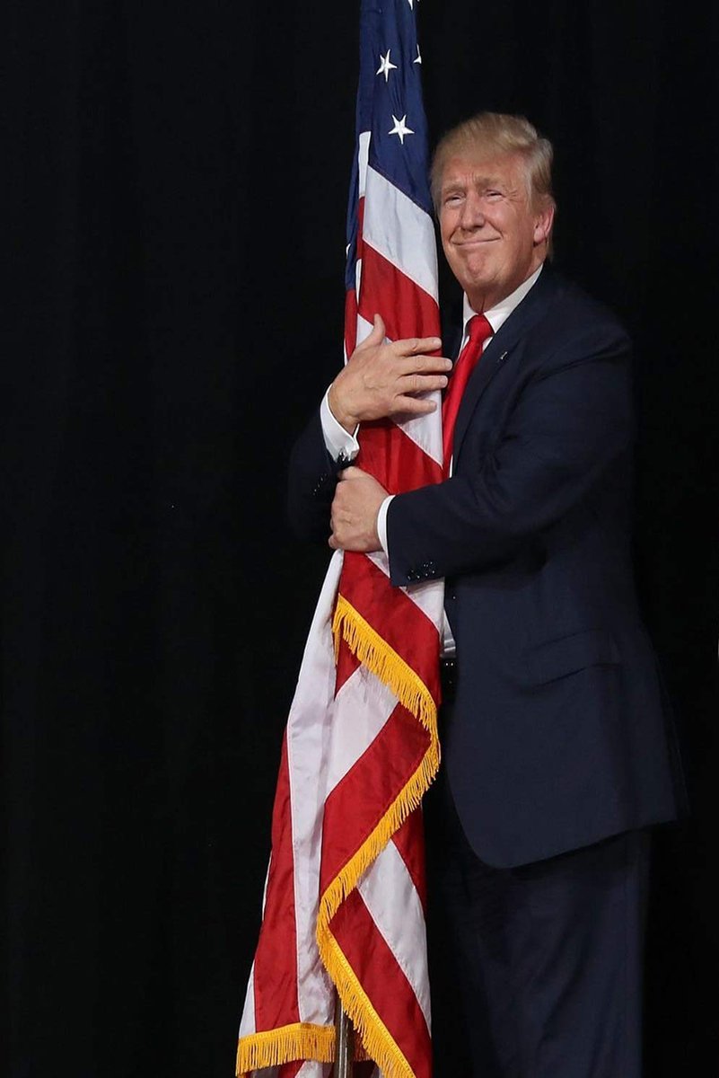 Donald Trump wall art print depicts him in a navy suit and red tie hugging a tasseled American flag against black.