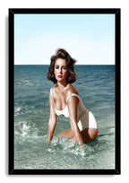 Elizabeth Taylor framed wall art print depicts the actress kneeling in blue-green surf in a white swimsuit, set in a black frame.