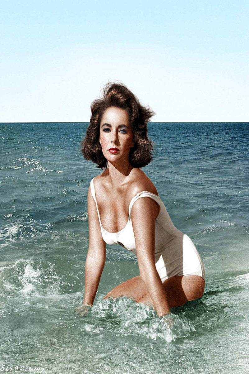 Elizabeth Taylor wall art print portrays the actress kneeling in surf in a white swimsuit beneath a pale blue sky.