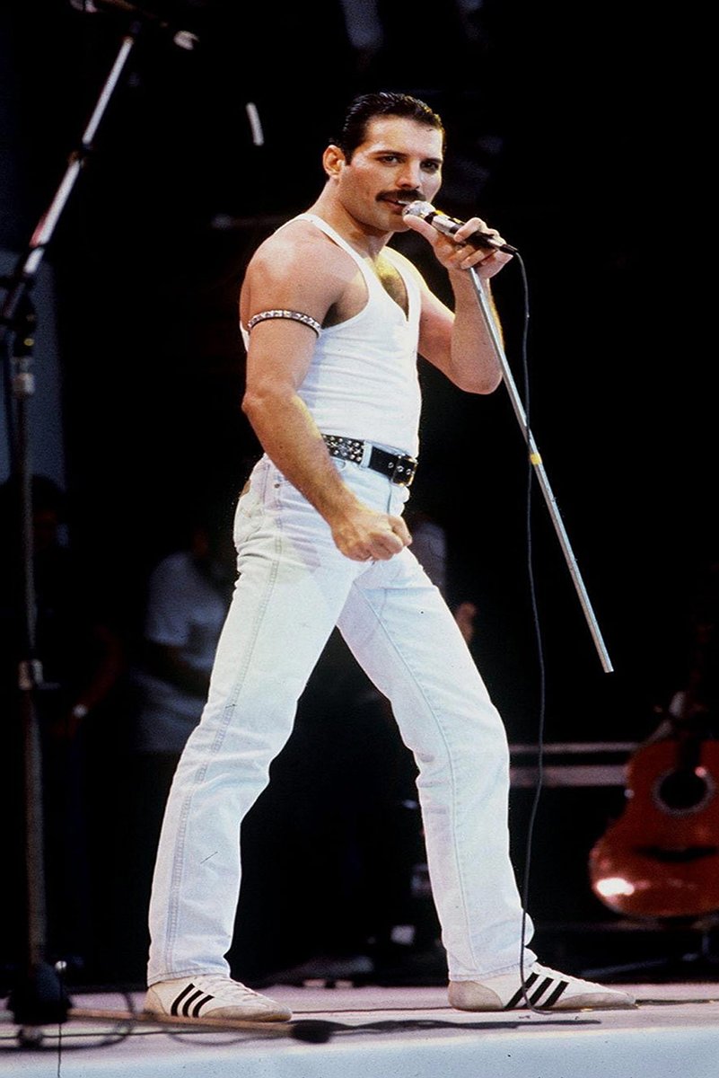 Freddie Mercury wall art print captures the singer on a dark stage in a white tank top and trousers, gripping a microphone.
