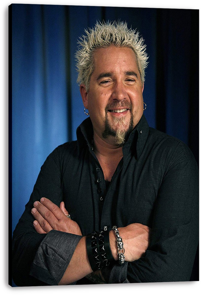 Guy Fieri canvas wall art print presents his smiling, arms-crossed portrait in a black shirt against deep blue curtains.