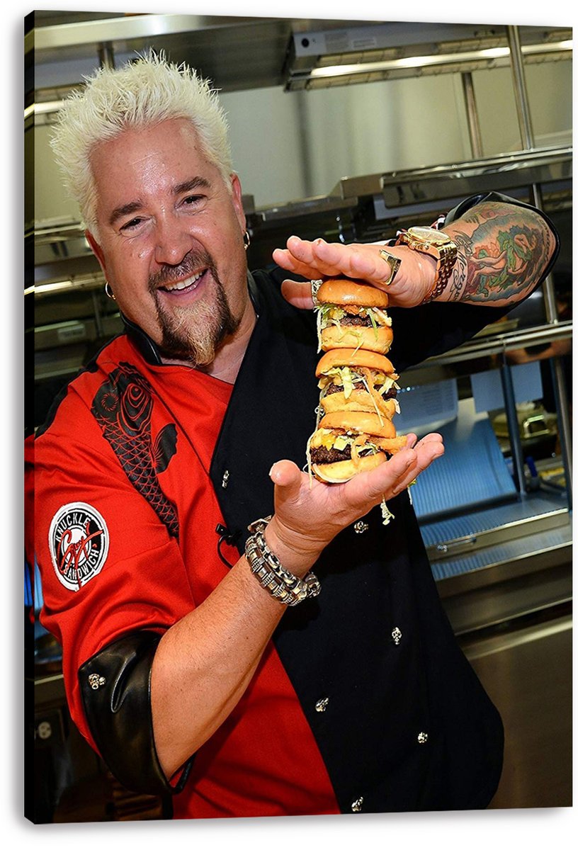 Guy Fieri canvas wall art print displays the tattooed chef holding a tall layered burger stack against stainless steel kitchen equipment.