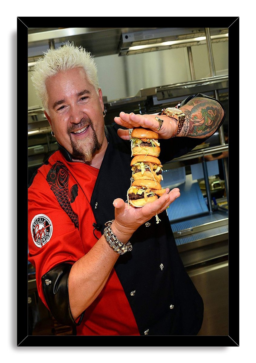 Guy Fieri framed wall art print presents the smiling chef, red shirt and black vest, balancing stacked burgers inside a black frame.