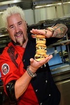 Guy Fieri poster features the smiling chef in a red and black shirt holding a towering stack of cheeseburgers in a commercial kitchen.