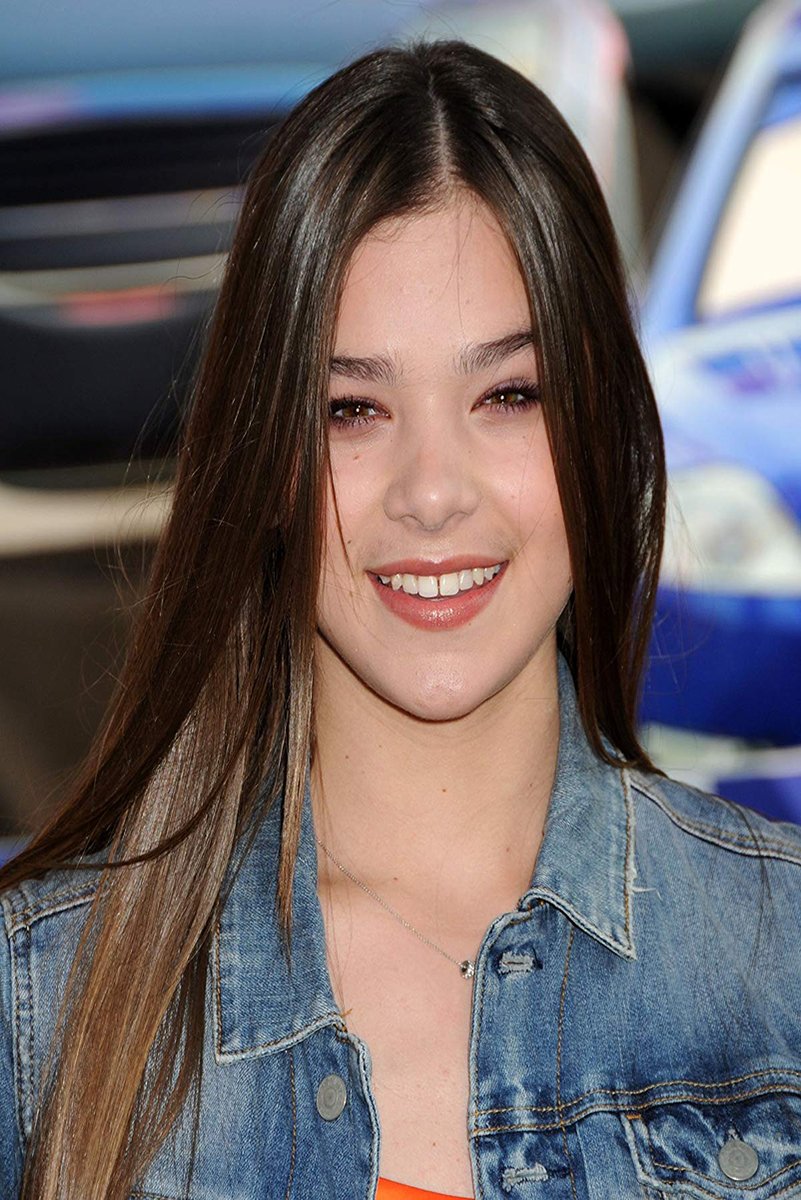 Hailee Steinfeld poster portrait with long dark hair, a warm smile, denim jacket, and blurred blue cars behind her.