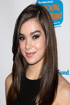 Hailee Steinfeld poster portrait with long brown hair, smoky eye makeup, and a black sleeveless top before a white event backdrop.