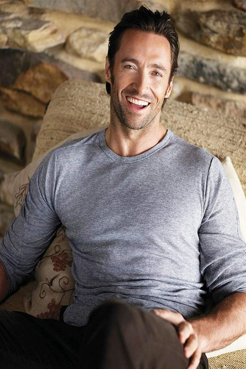 Hugh Jackman poster portrait with the actor smiling on a patterned sofa in a gray long-sleeve shirt before a stone wall.