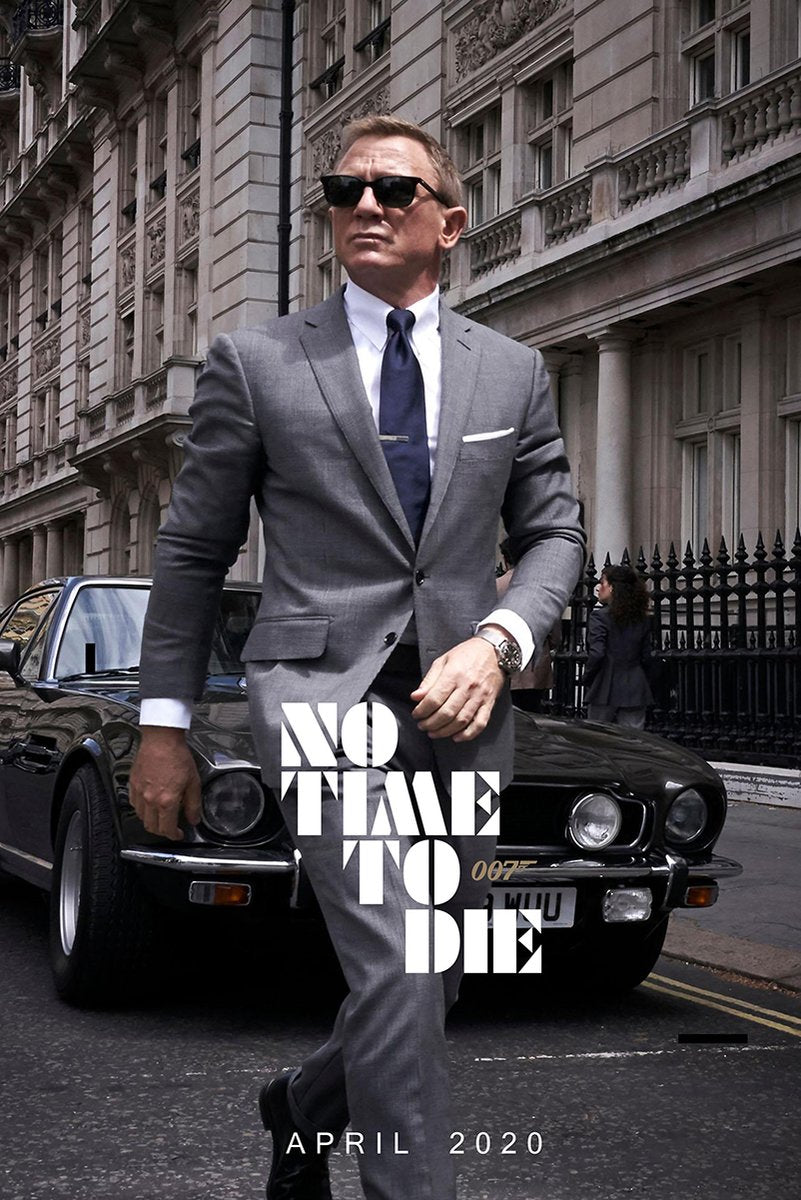 James Bond No Time To Die poster features Daniel Craig in a gray suit and sunglasses walking before a black sports car on a city street.