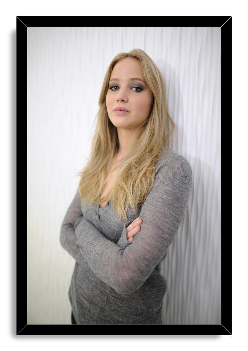 Jennifer Lawrence framed wall art print presents a blonde woman in gray knitwear with folded arms, set against white drapery in a black frame.
