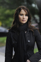 Jennifer Lawrence poster portrait with the actress in a black trench coat and scarf against a softly blurred green outdoor background.