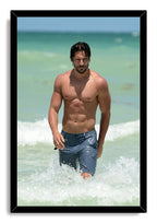Joe Manganiello framed wall art print shows a shirtless man in blue shorts walking through breaking ocean waves.