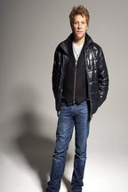 Jon Bon Jovi wall art print features a full-length studio portrait in a black puffer jacket, dark vest, and blue jeans.