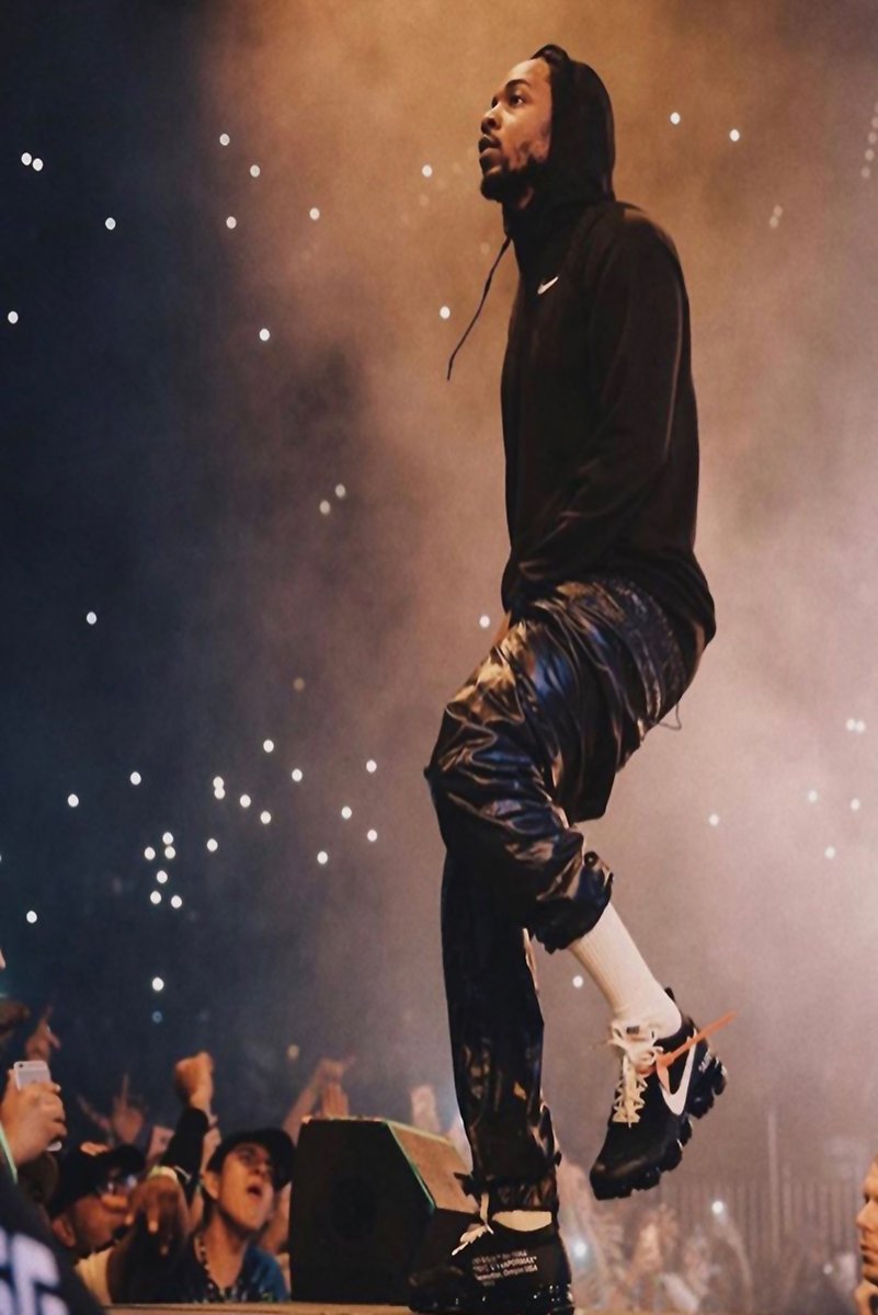 Kendrick Lamar poster captures the rapper onstage in a black hoodie and glossy pants amid warm concert haze and crowd lights.