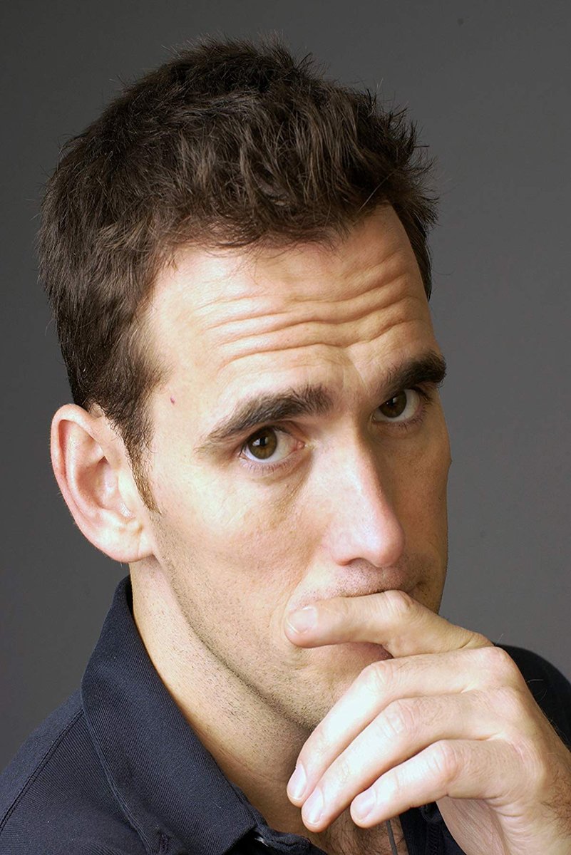 Matt Dillon poster portrait with tousled dark hair, a contemplative hand-to-mouth pose, and a charcoal gray background.
