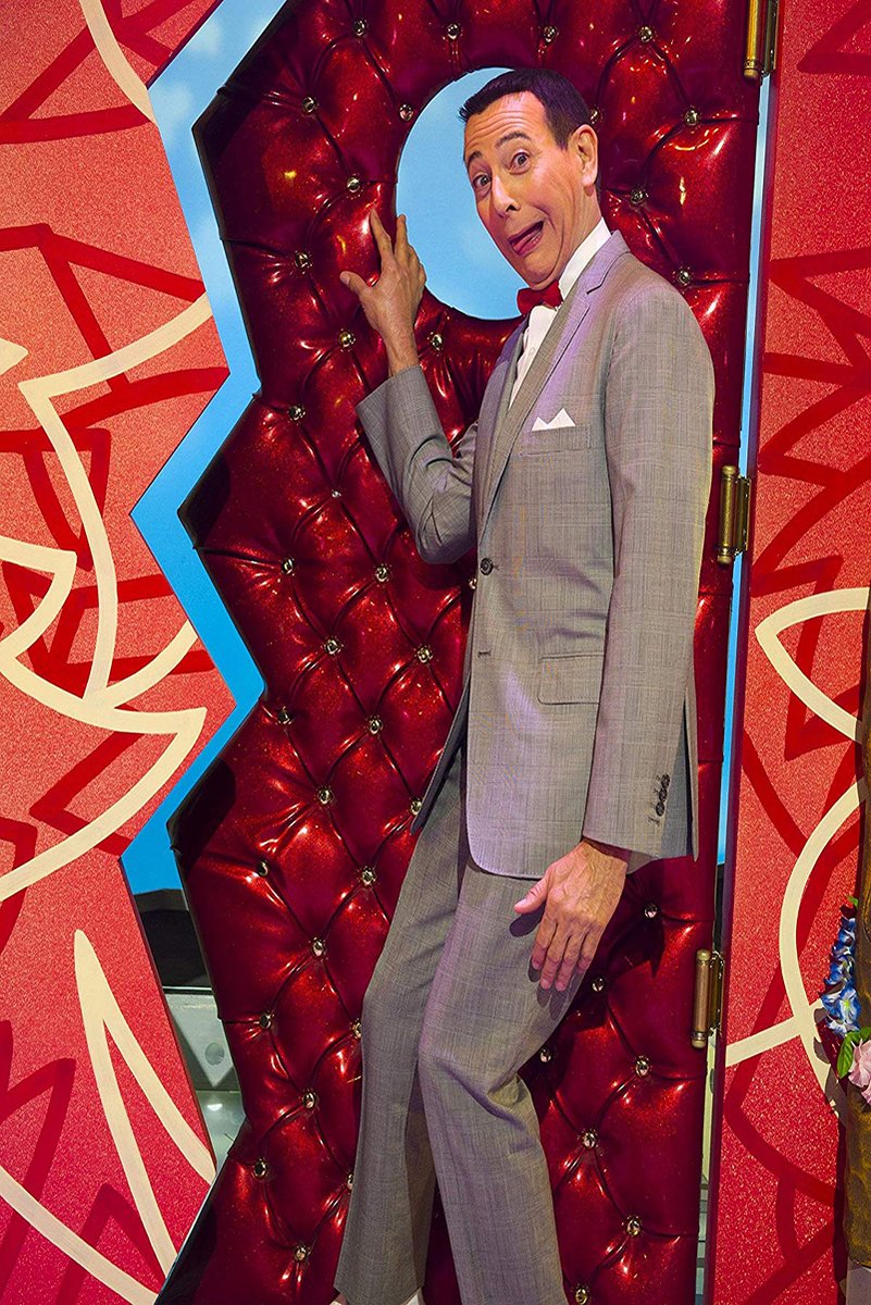 Pee-wee Herman poster depicts him in a gray suit and red bow tie beside a glossy red tufted doorway.