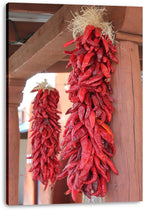 Red chili ristras canvas wall art print with vivid dried peppers and straw ties against warm adobe-toned wood.