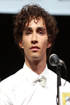 Robert Sheehan poster portrait in a white tuxedo and bow tie, standing beside a microphone against a dark backdrop.