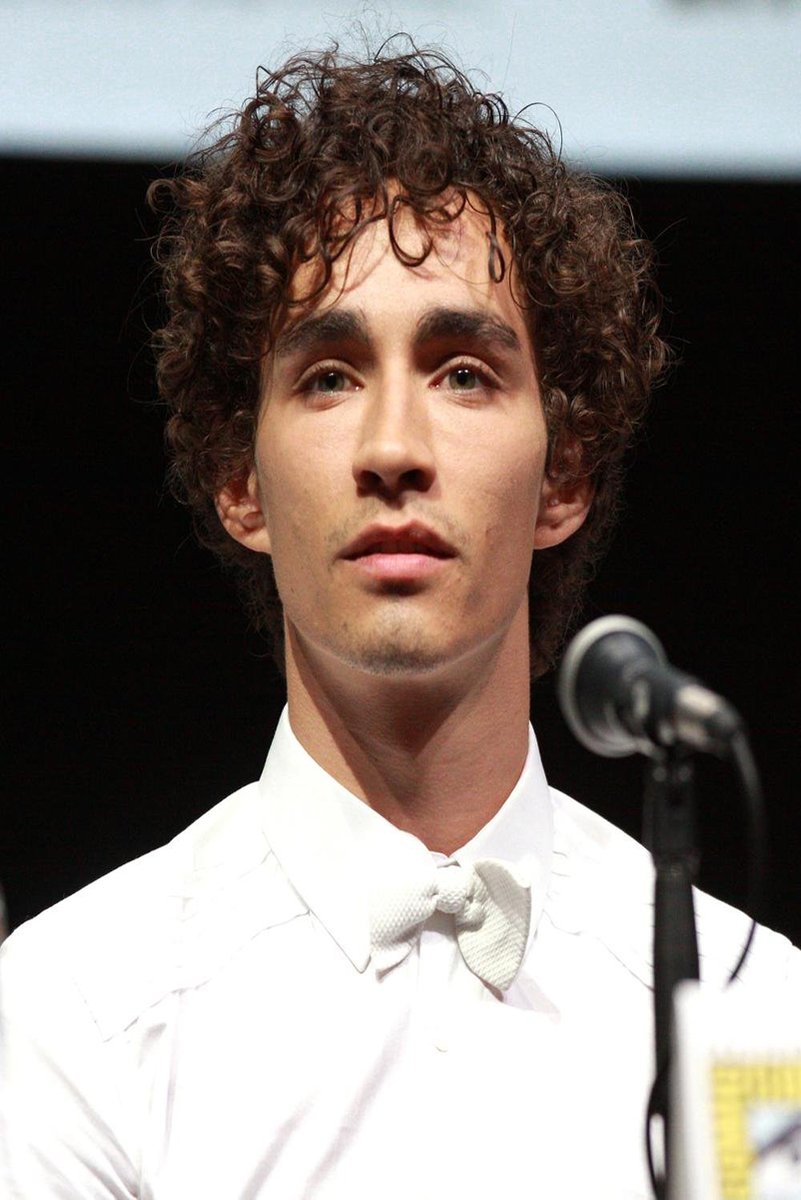 Robert Sheehan poster portrait in a white tuxedo and bow tie, standing beside a microphone against a dark backdrop.
