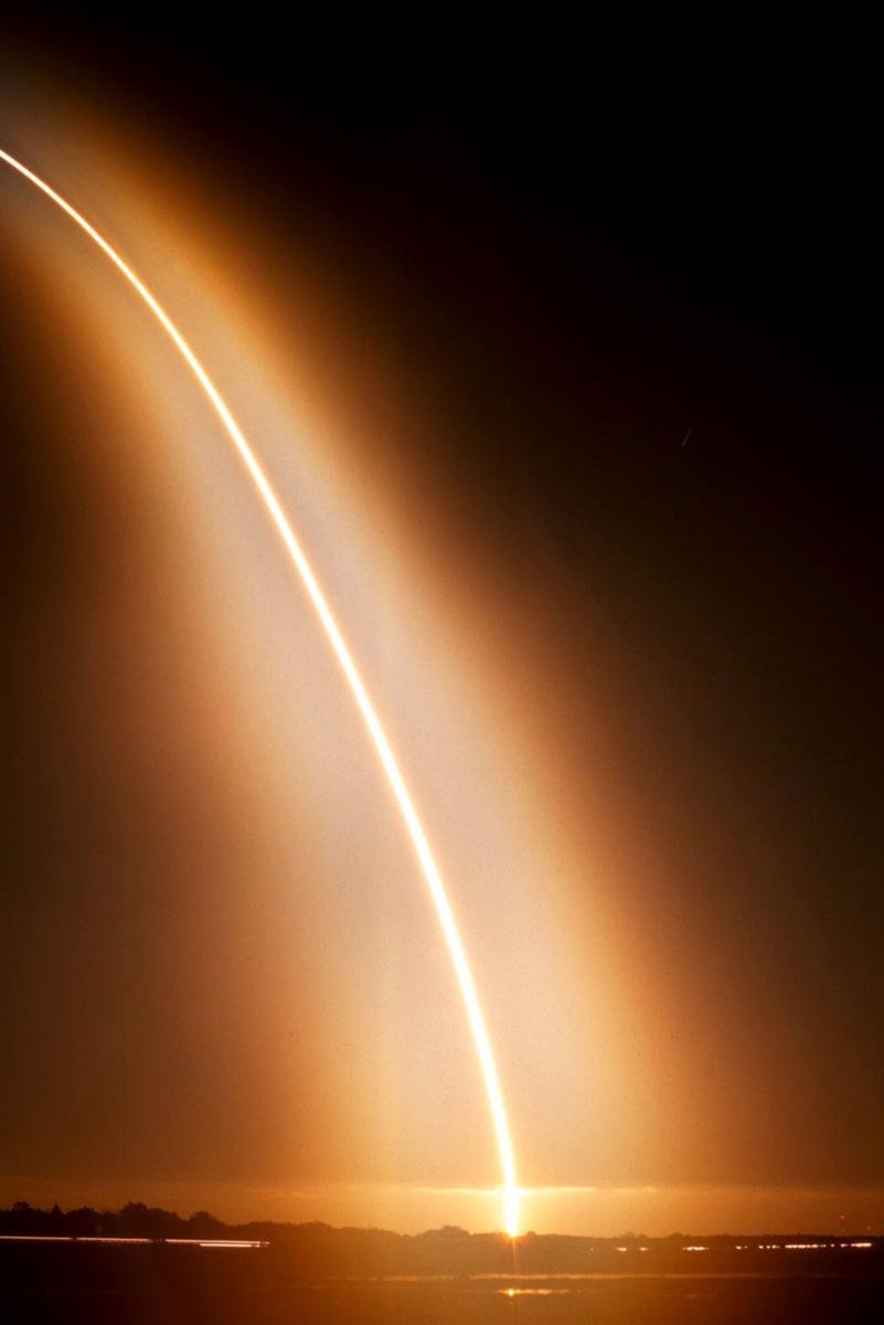 Rocket launch wall art print captures a sweeping white-orange light trail rising from a glowing horizon beneath a dark sky.