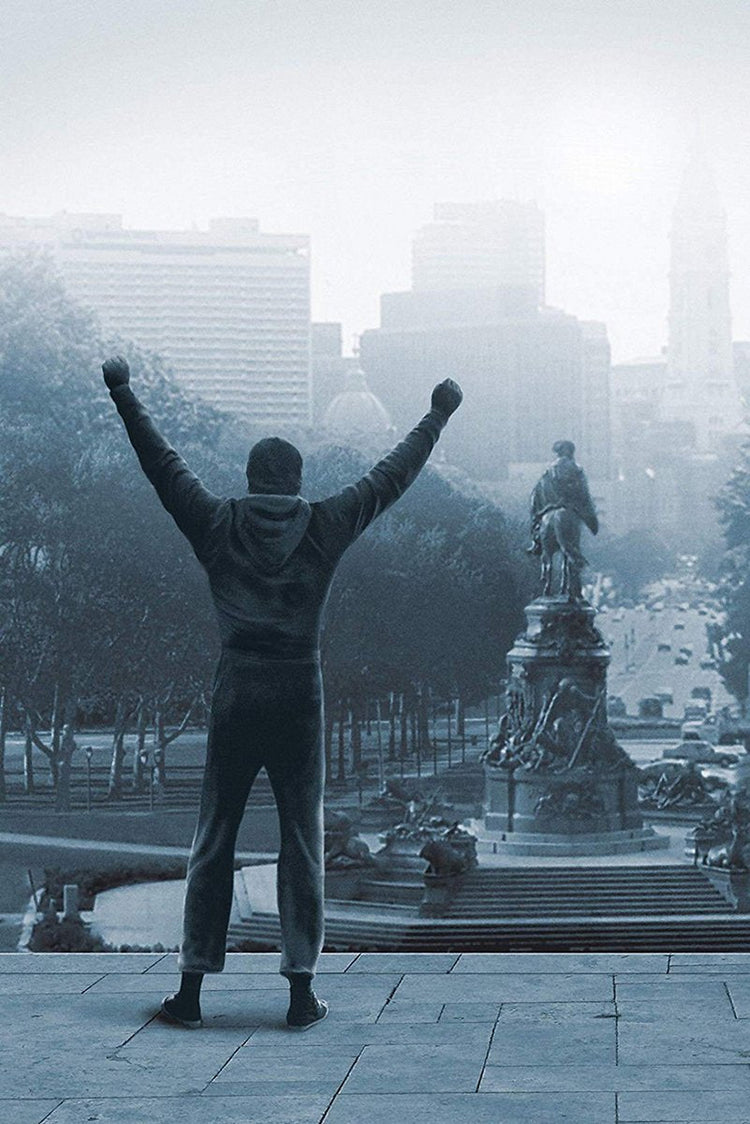 Rocky Balboa wall art print depicts a man from behind raising both fists atop stone steps above a misty city street.