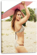 Ronda Rousey canvas wall art print displays her in a gray bikini lifting a pink surfboard against a sandy beach backdrop.