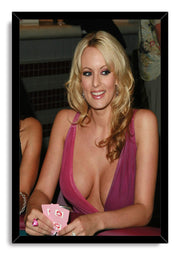 Stormy Daniels framed wall art print portrays her smiling while holding cards at a poker table, set in a black frame.