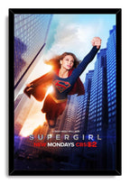 Supergirl framed wall art print in a black frame, featuring the heroine flying through a sunlit blue city skyline.