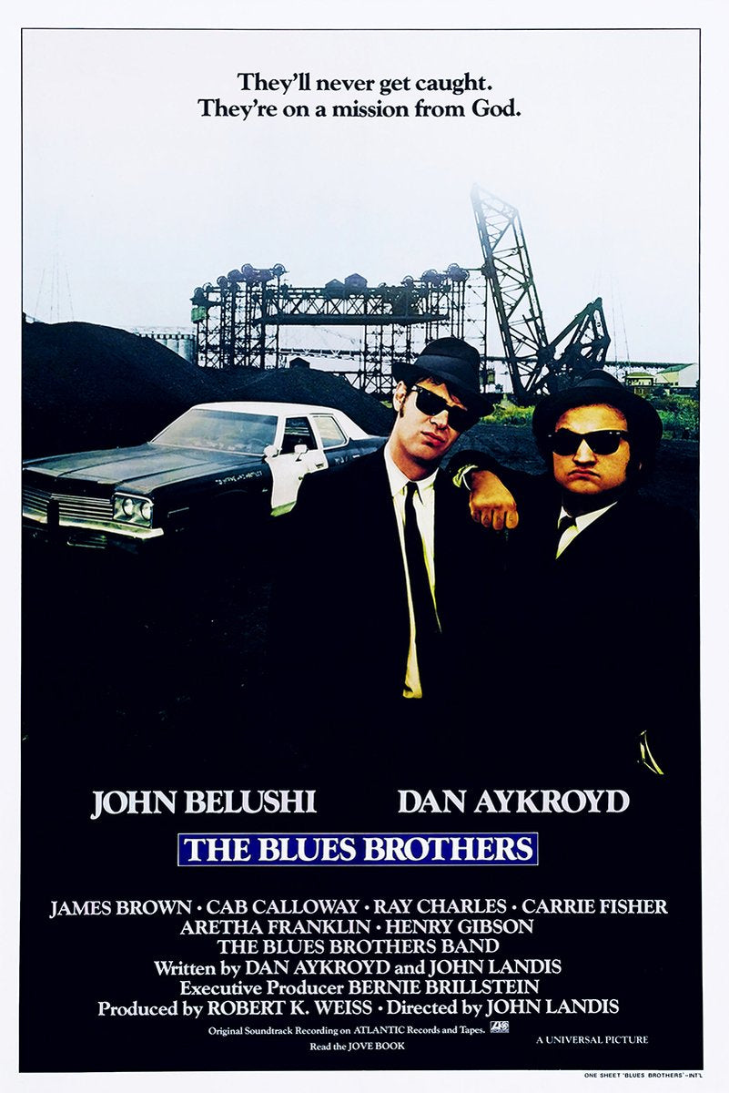 The Blues Brothers poster features two men in black suits, hats and sunglasses beside a police car in an industrial yard.