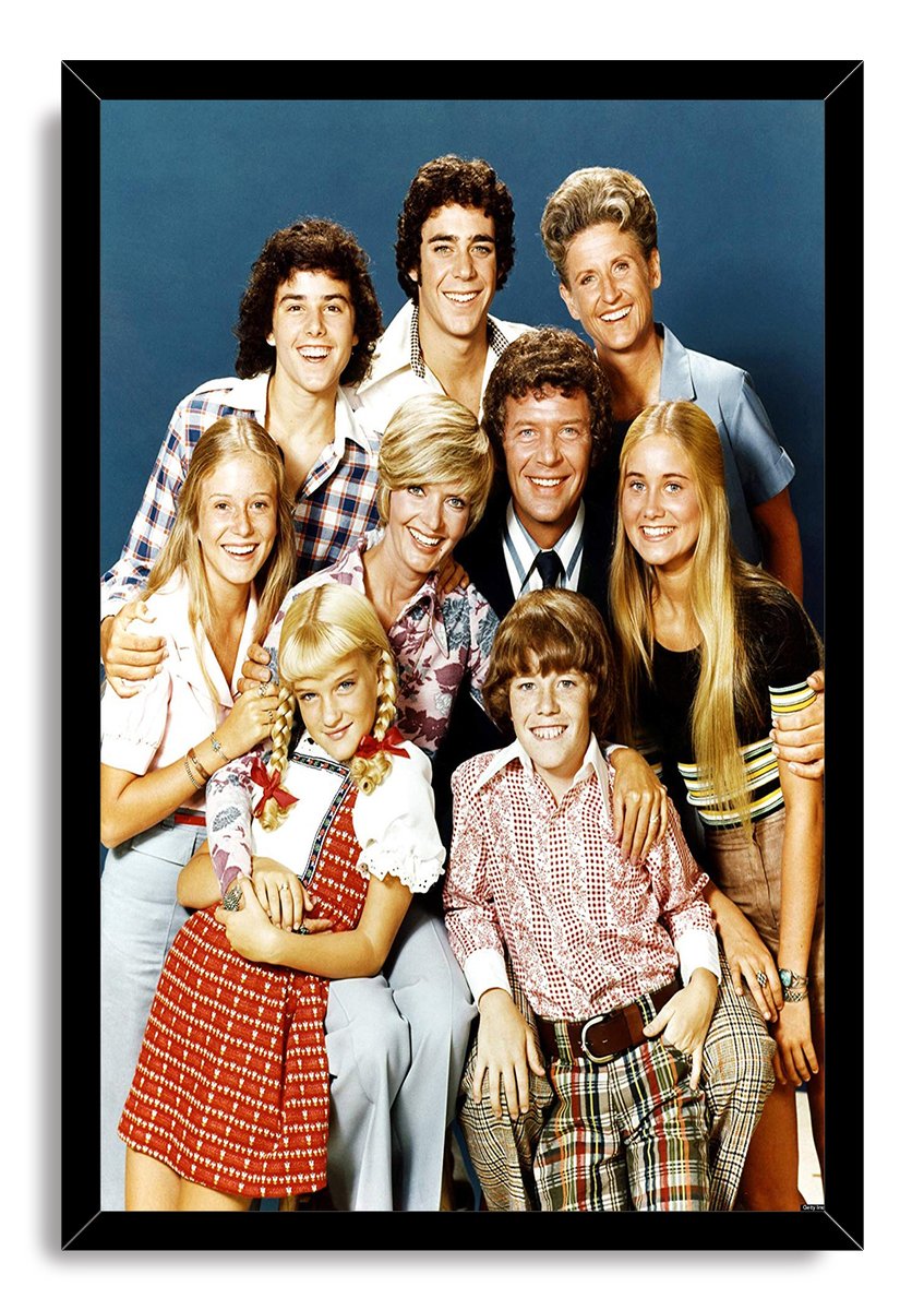 The Brady Bunch cast framed wall art print displays nine smiling actors in colorful retro outfits within a black mitered frame.