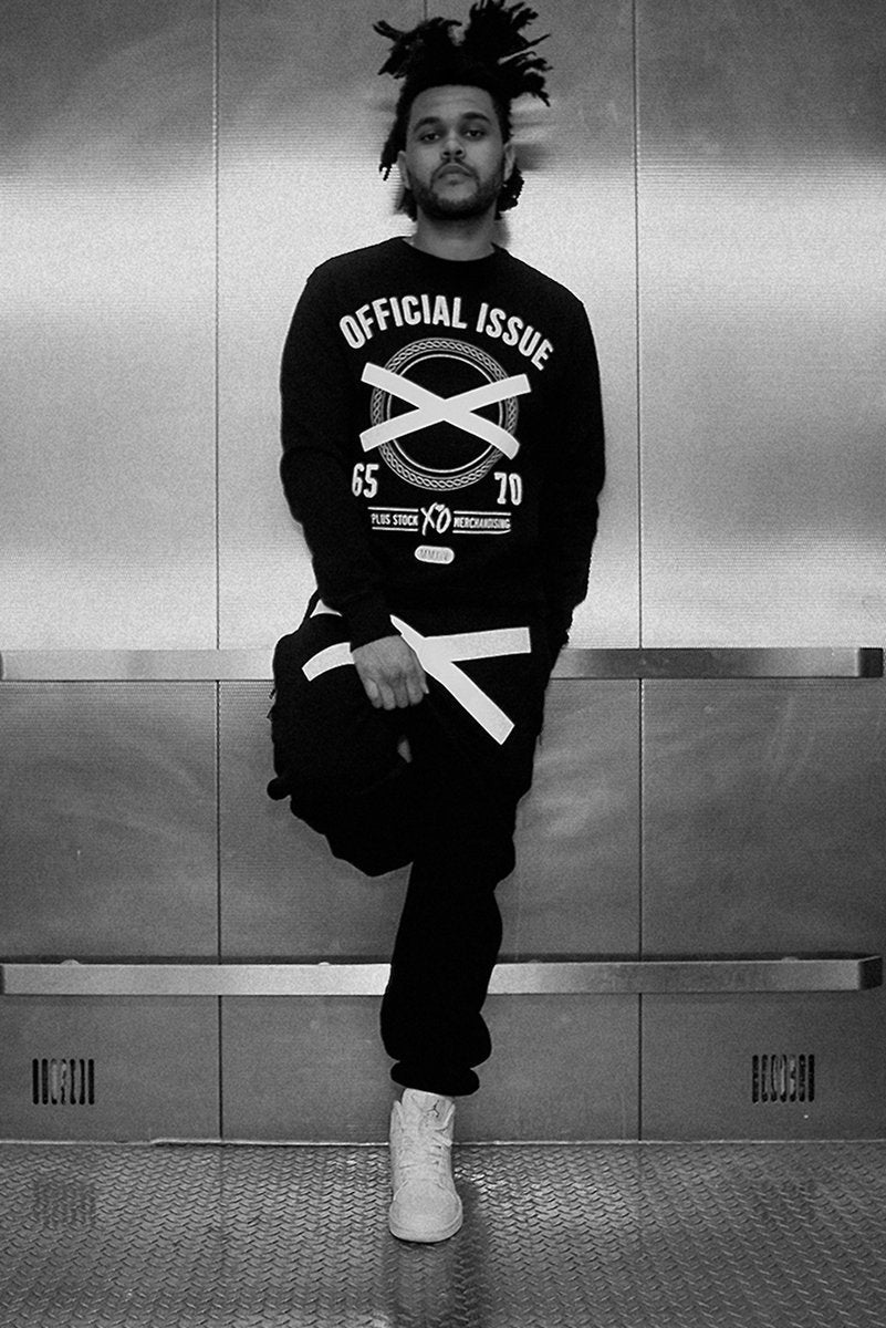 The Weeknd poster in black and white, standing in a metal elevator wearing a graphic sweatshirt and white sneakers.