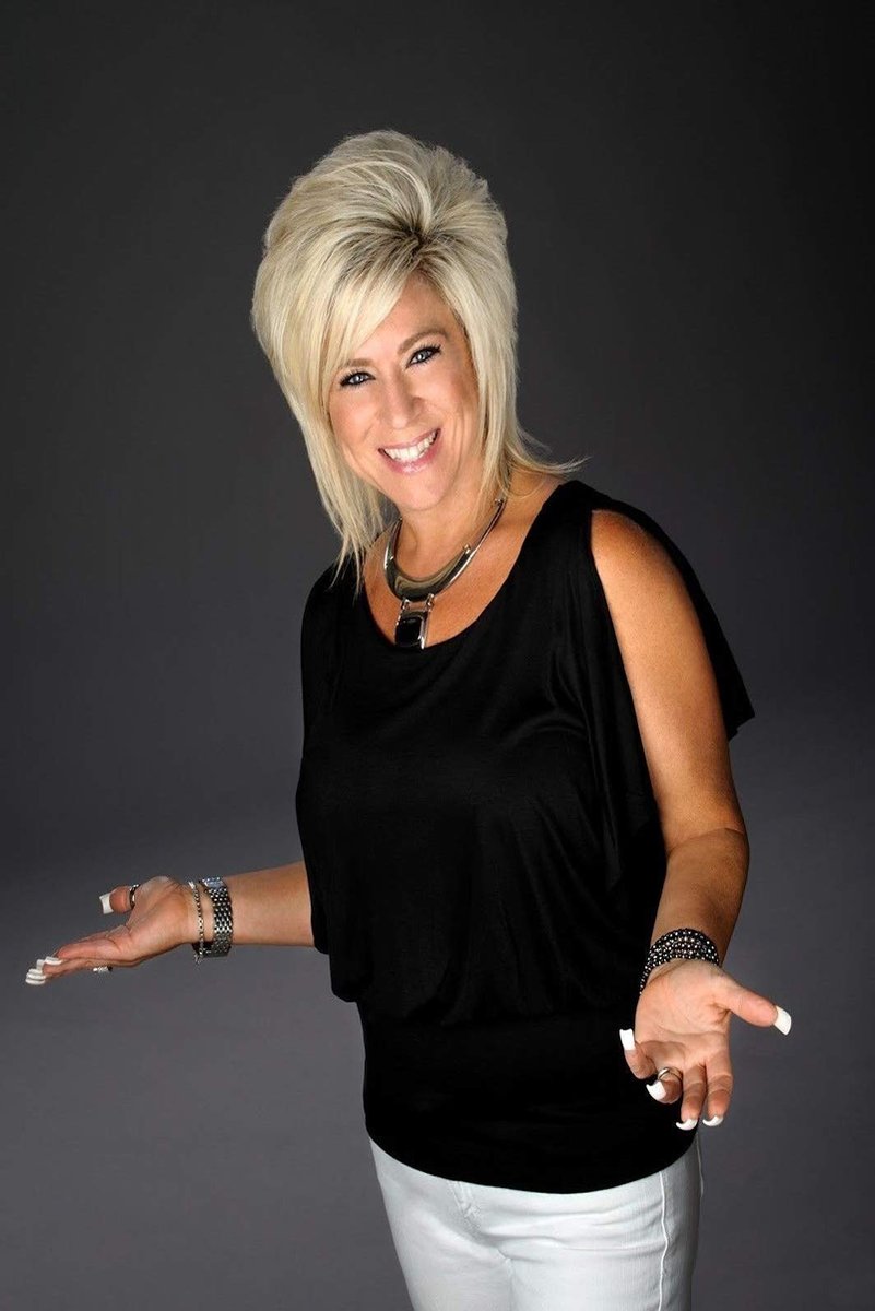 Theresa Caputo poster portrait with blonde hair, a black top and white pants, smiling with open hands against gray.