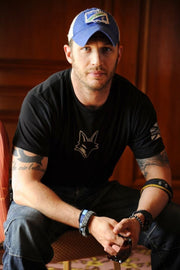 Tom Hardy poster portrait of the actor seated in a blue cap and black T-shirt, holding sunglasses against dark wood paneling.