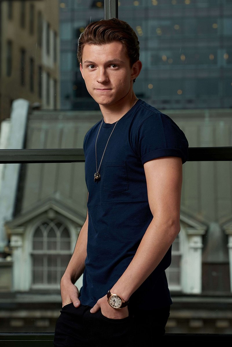 Tom Holland poster portrait in a navy T-shirt and pendant necklace, standing before large windows with hands in pockets.